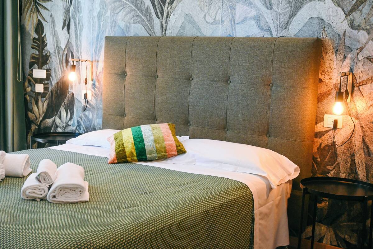 A neatly made bed with a green and white bedspread, a striped pillow, and rolled towels on the side. There are wall-mounted lights and a decorative wall mural.