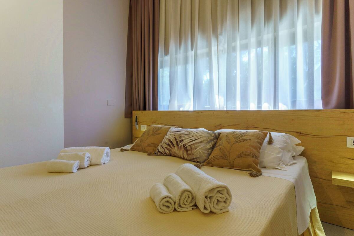 A neatly made bed with rolled towels, a wooden headboard, and a window with sheer curtains.