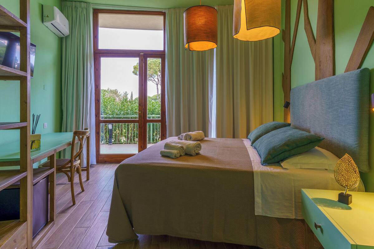 A bedroom with a bed, a desk, a chair, a bedside table, and a window with curtains.
