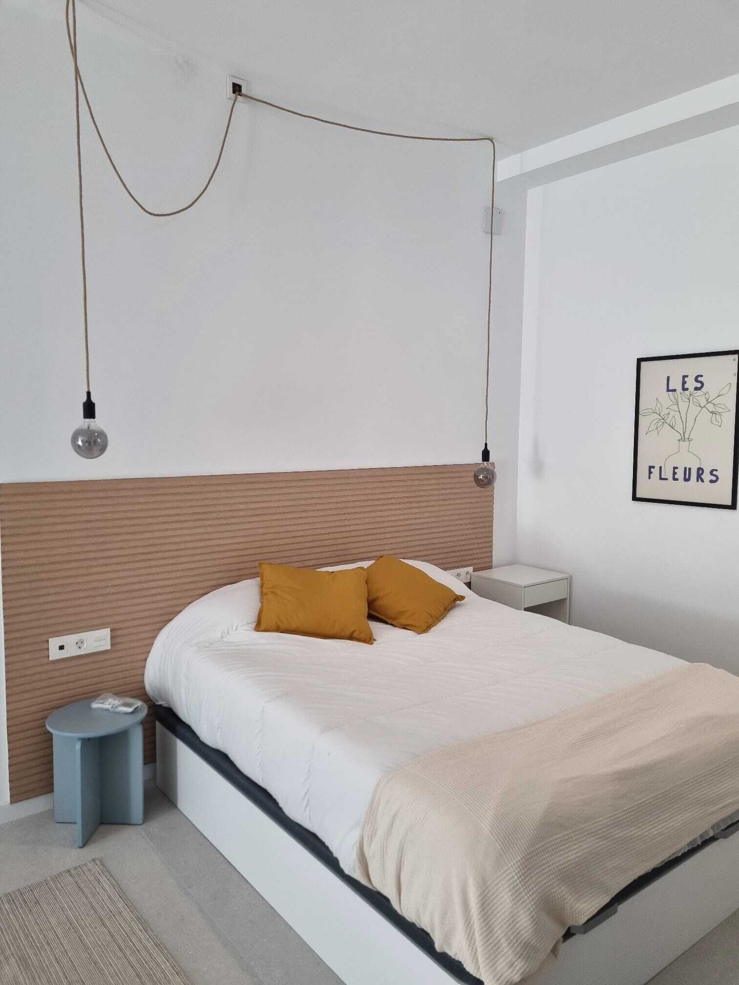 Un dormitorio moderno con cama blanca, almohadas color mostaza, cabecero de madera, una mesita de noche y una lámpara de pared, además de un cuadro enmarcado.