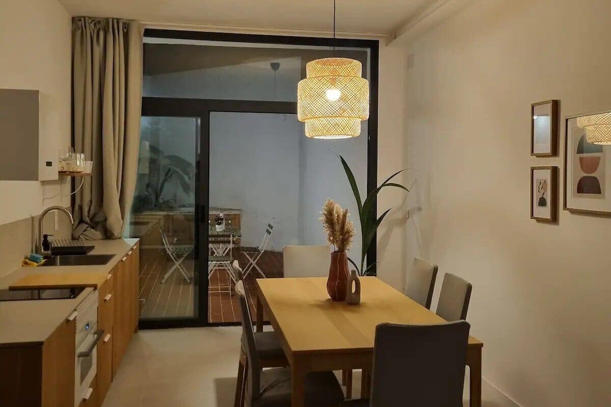 Un espace repas moderne avec une table et des chaises en bois, un luminaire suspendu et une vue sur une terrasse extérieure, accessible par une porte vitrée.