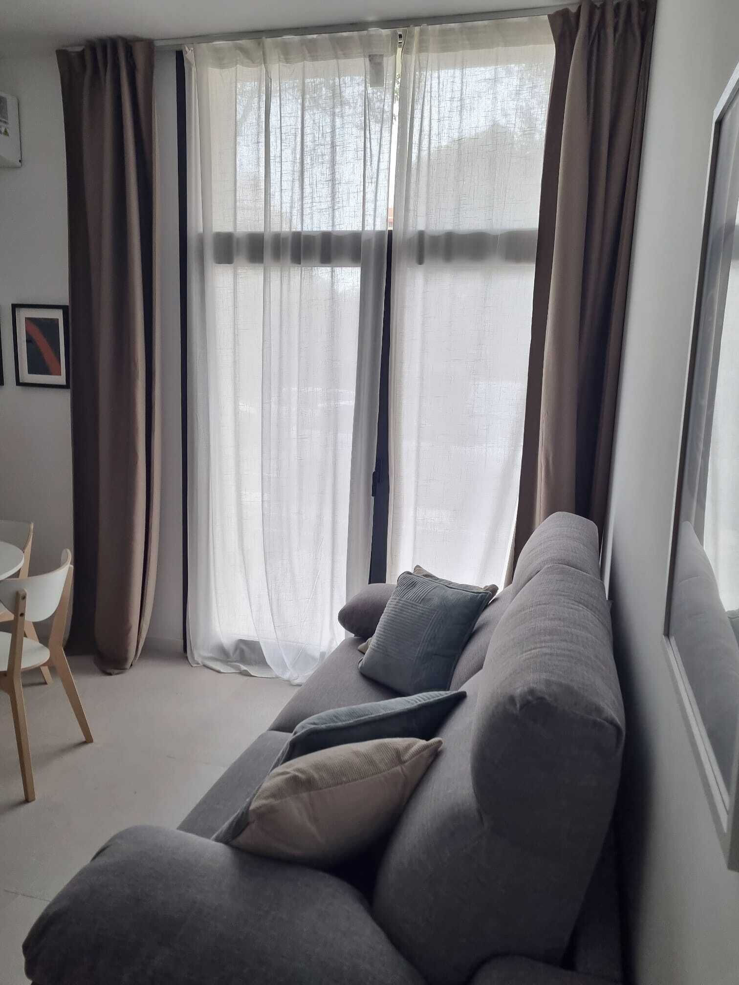 A living room with a grey sofa, a small table, and a window with sheer curtains.