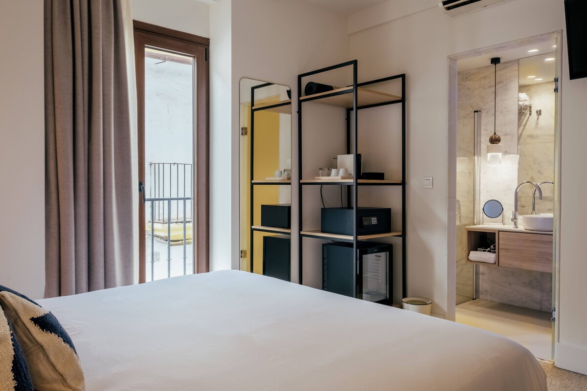 Habitación de hotel con cama, armario, baño con lavabo y espejo, y vistas al balcón.