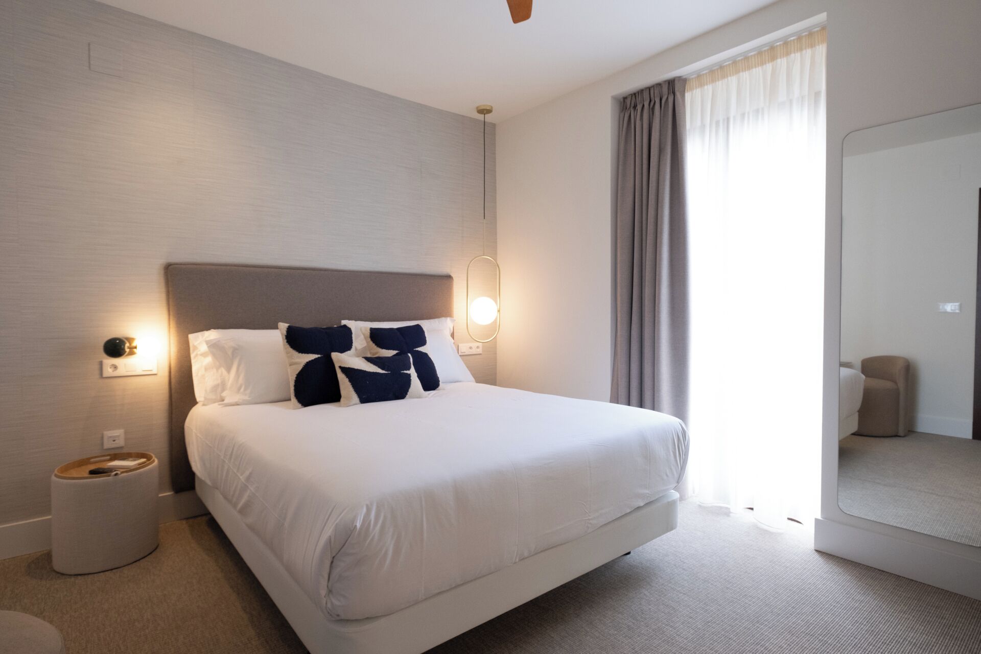 Una habitación de hotel moderna con una cama grande, una mesita de noche, una lámpara y vistas al exterior a través de una ventana con cortinas.