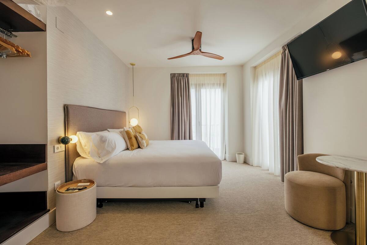 Une chambre d’hôtel moderne avec un grand lit, un téléviseur à écran plat fixé au mur, un ventilateur de plafond et une petite table ronde.