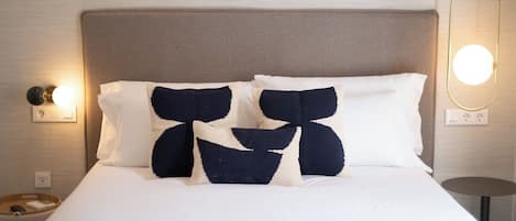 Een net opgemaakt bed met witte lakens en twee decoratieve kussens met een geometrisch patroon. Aan weerszijden van het bed hangen wandlampen.