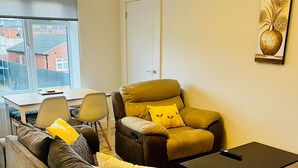 Ein modernes Wohnzimmer mit Sofa, Sessel und Couchtisch. Es gibt einen Esstisch mit Stühlen, eine Tür und ein Fenster mit Blick auf Gebäude.