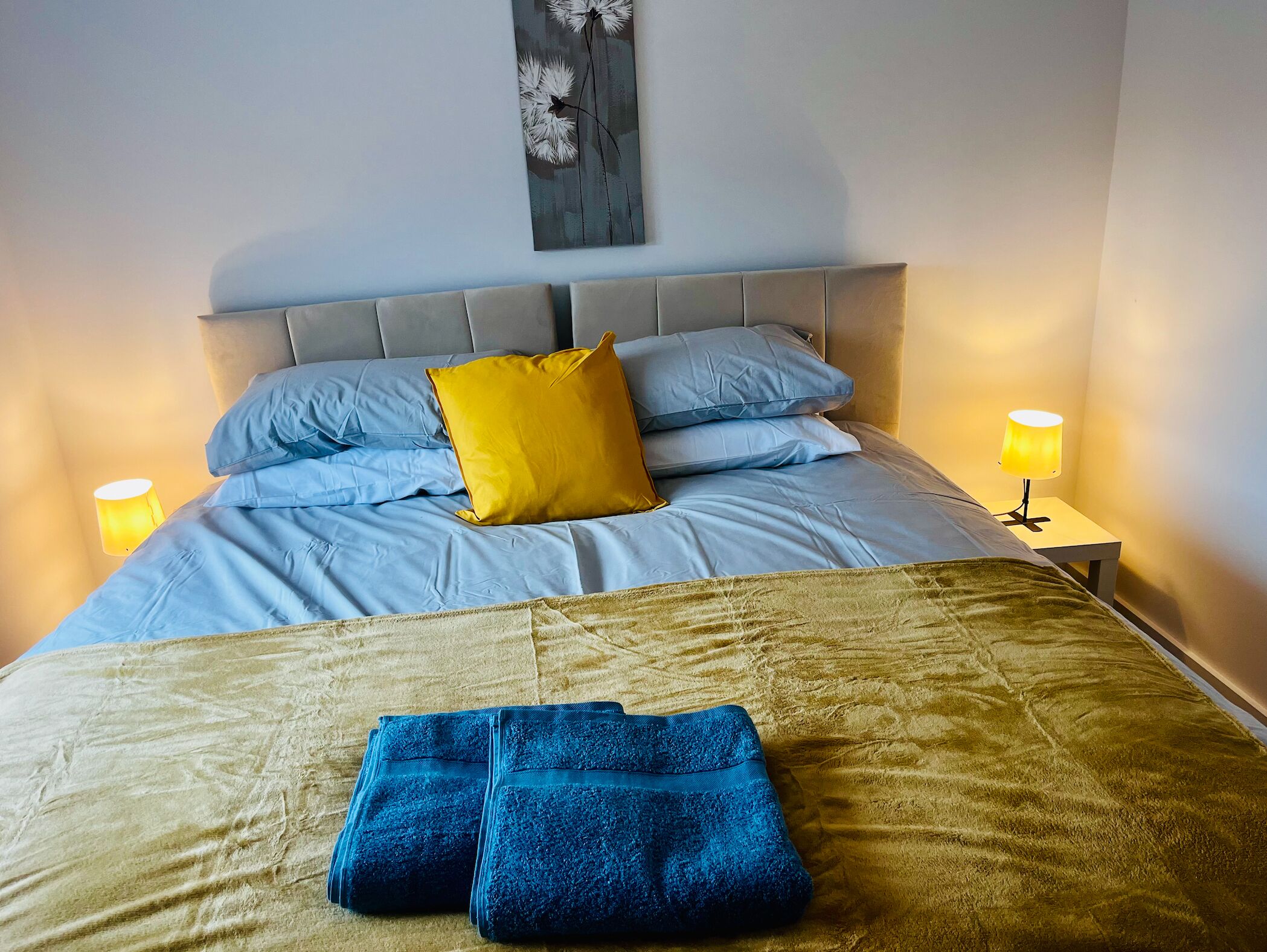 A neatly made bed with blue and yellow pillows, a grey headboard, and two bedside lamps.