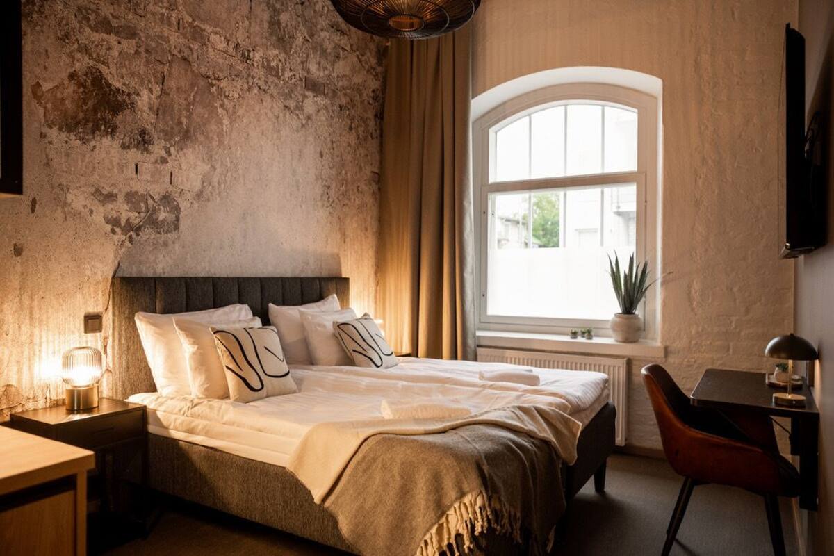 A bedroom with a large bed, a desk, a chair, a window with curtains, and a plant.