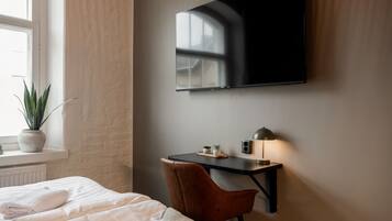 A hotel room with a bed, a desk, a chair, a TV mounted on the wall, and a window with a plant on the sill.