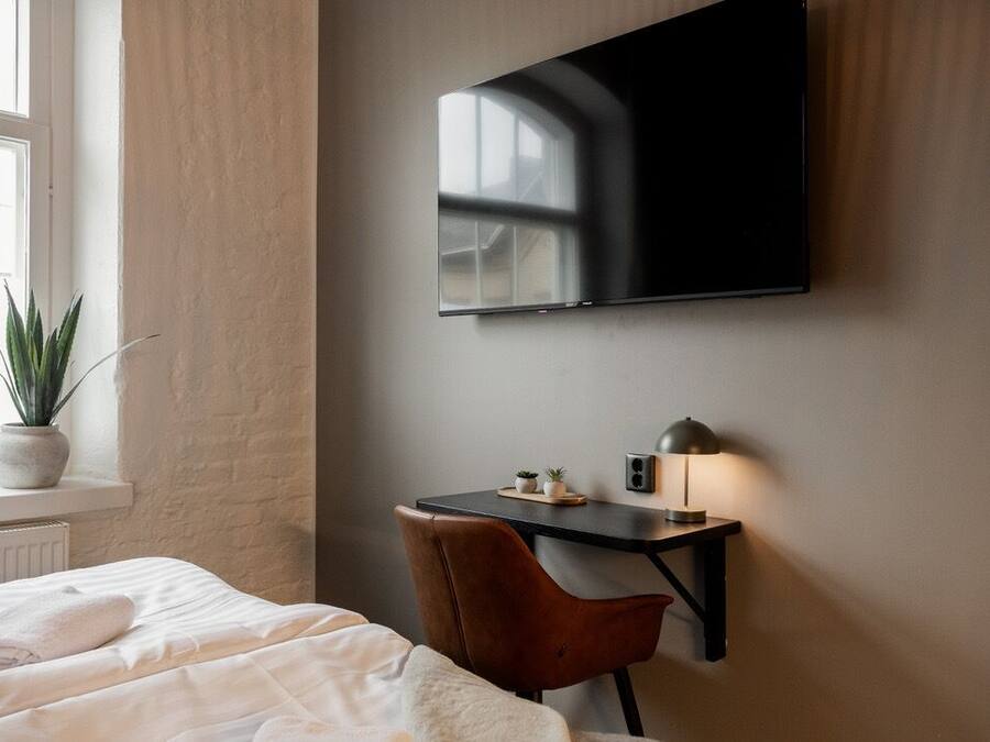 A hotel room with a bed, a desk, a chair, a TV mounted on the wall, and a window with a plant on the sill.