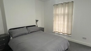 Ein Schlafzimmer mit Bett, Nachttisch, Lampe und einem Fenster mit Vorhängen.