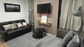 Ein modernes Wohnzimmer mit einer schwarzen Ledersofa, einem runden Glastisch und einem grauen Sessel. Ein Flachbildfernseher ist an einer Holzvertäfelung befestigt, und auf beiden Seiten des Fernsehers stehen zwei Lampen.