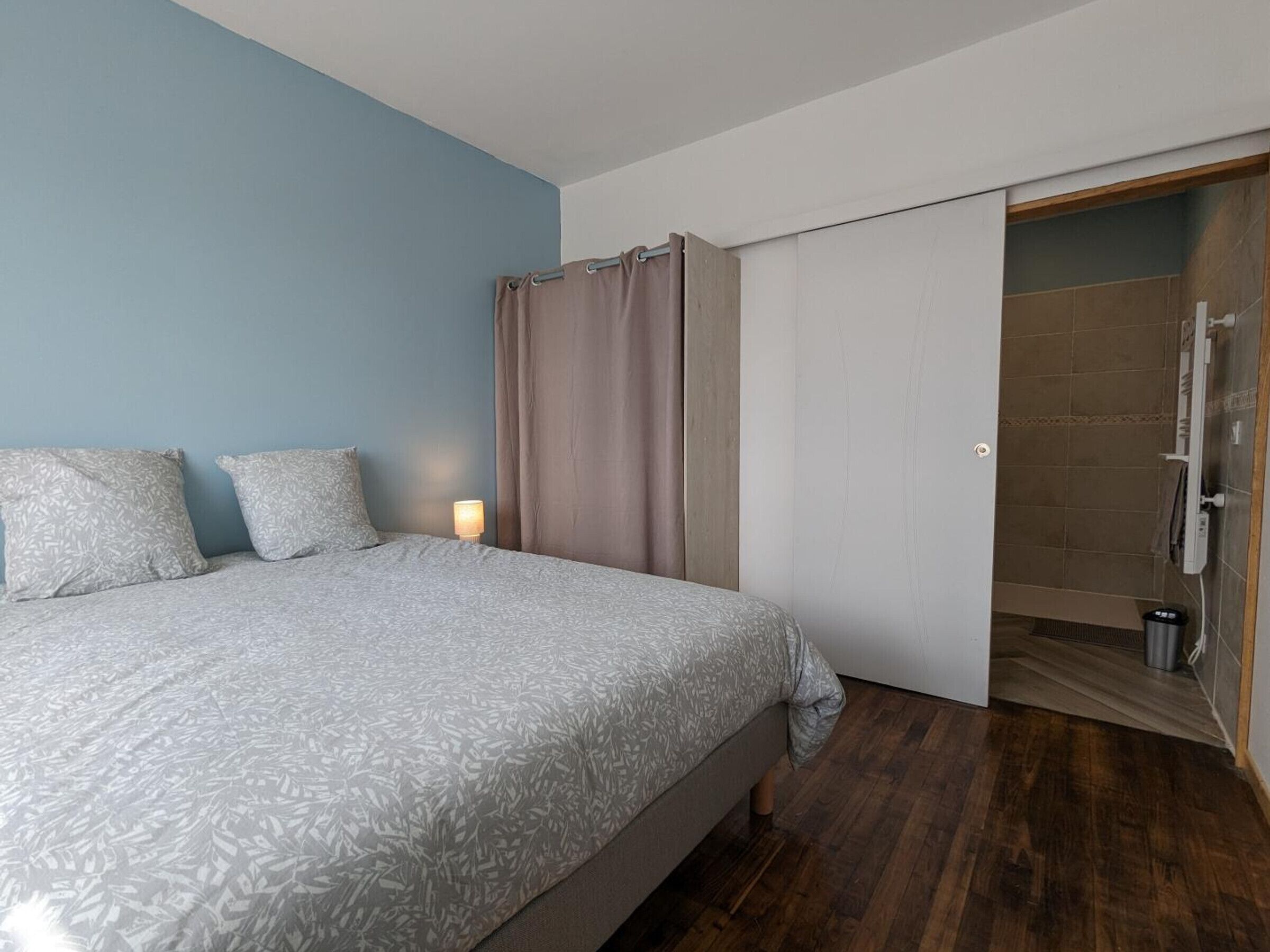 A bedroom with a bed, a nightstand, a lamp, and a shower area.