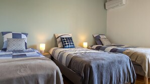 Ein Zimmer mit drei nebeneinander stehenden Betten, jedes mit einer anderen Bettdecke. An der Wand befinden sich ein Deckenventilator und eine Klimaanlage.