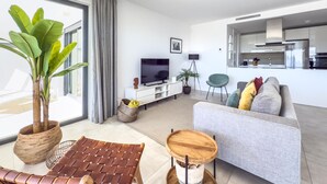 A modern living room with a sofa, coffee table, and a TV. There is a large window with curtains, a potted plant, and a kitchen area visible in the background. - Modern apartment with terrace, pool, and Wi-Fi in Estepona (Estepona)