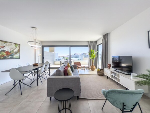 A modern living room with a sofa, dining table, chairs, a TV, and a large window offering a city view. - Modern apartment with terrace, pool, and Wi-Fi in Estepona (Estepona)