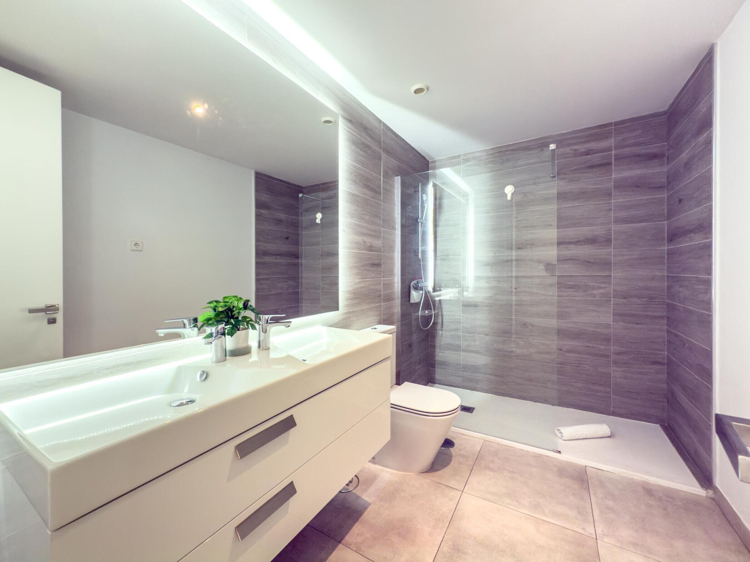 A modern bathroom with a large mirror, dual sinks, a toilet, and a walk-in shower.