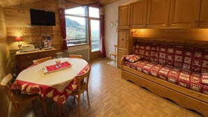 A cozy room with a wooden ceiling, a dining table with a red tablecloth, a sofa with a patterned cover, and a window offering a view of the outdoors. - Spacious 2-room apartment for 5 people in Chinaillon (Le Grand-Bornand)