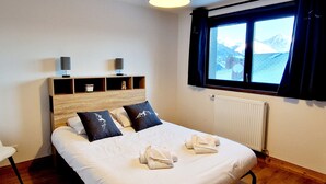 Una habitación de hotel con una cama grande, un cabecero de madera, una mesita de noche con una lámpara, una ventana con vistas a montañas y un radiador.