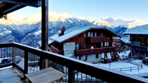 Un chalet de madera con base de piedra, un balcón y vistas a montañas nevadas.