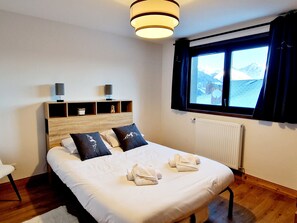 Una habitación de hotel con una cama grande, un cabecero de madera, una mesita de noche con una lámpara, una ventana con vistas a montañas y un radiador.