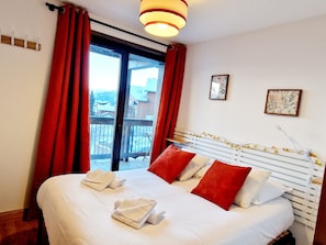 A hotel room with a bed, red curtains, a balcony, and two framed pictures on the wall. - Charming apartment in Montalbert center with balcony and parking, close to slopes (La Plagne Montalbert)