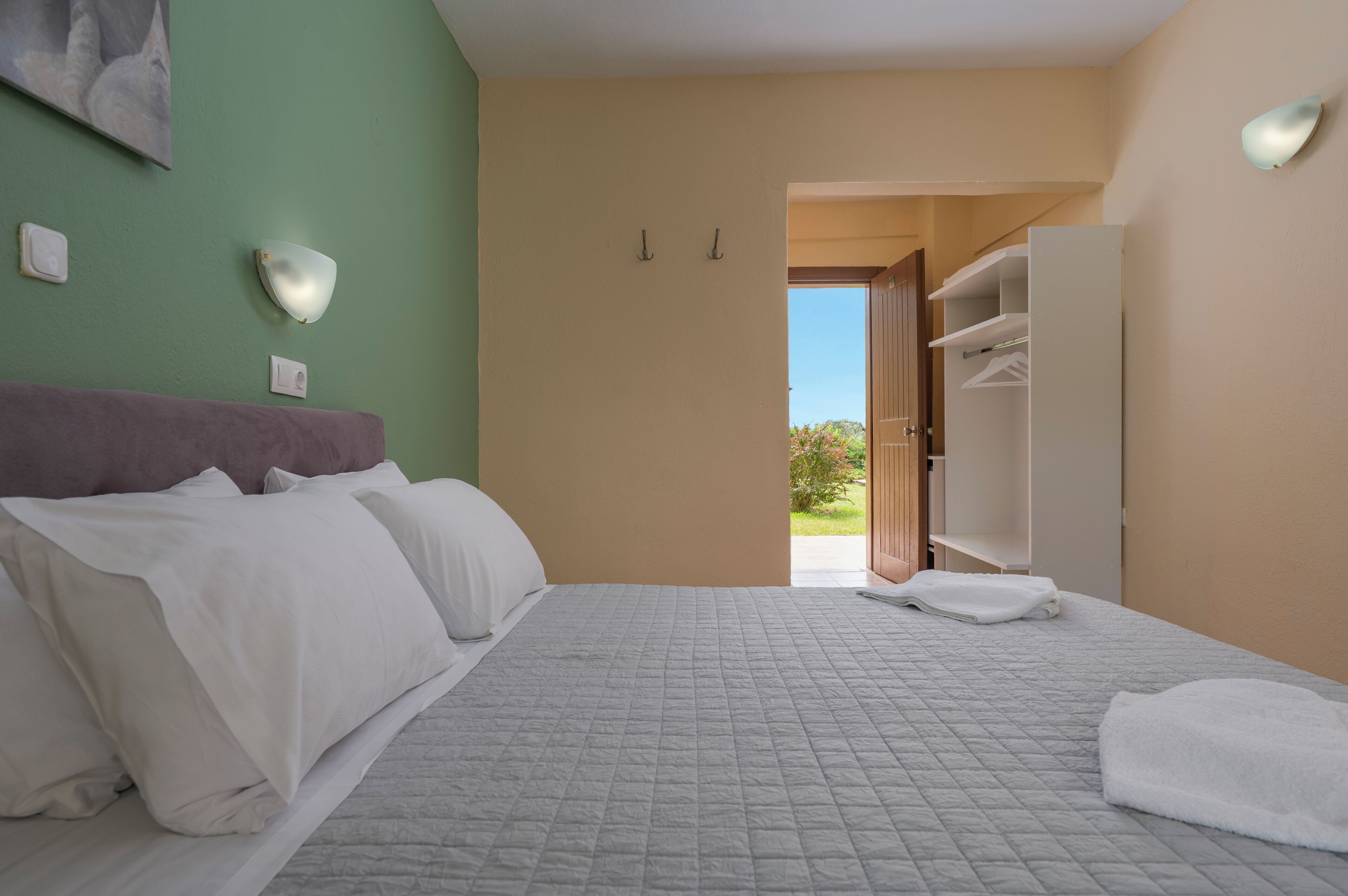 A hotel room with a bed, a nightstand, a wall-mounted light, a door leading to a balcony, and a view of the outdoors.