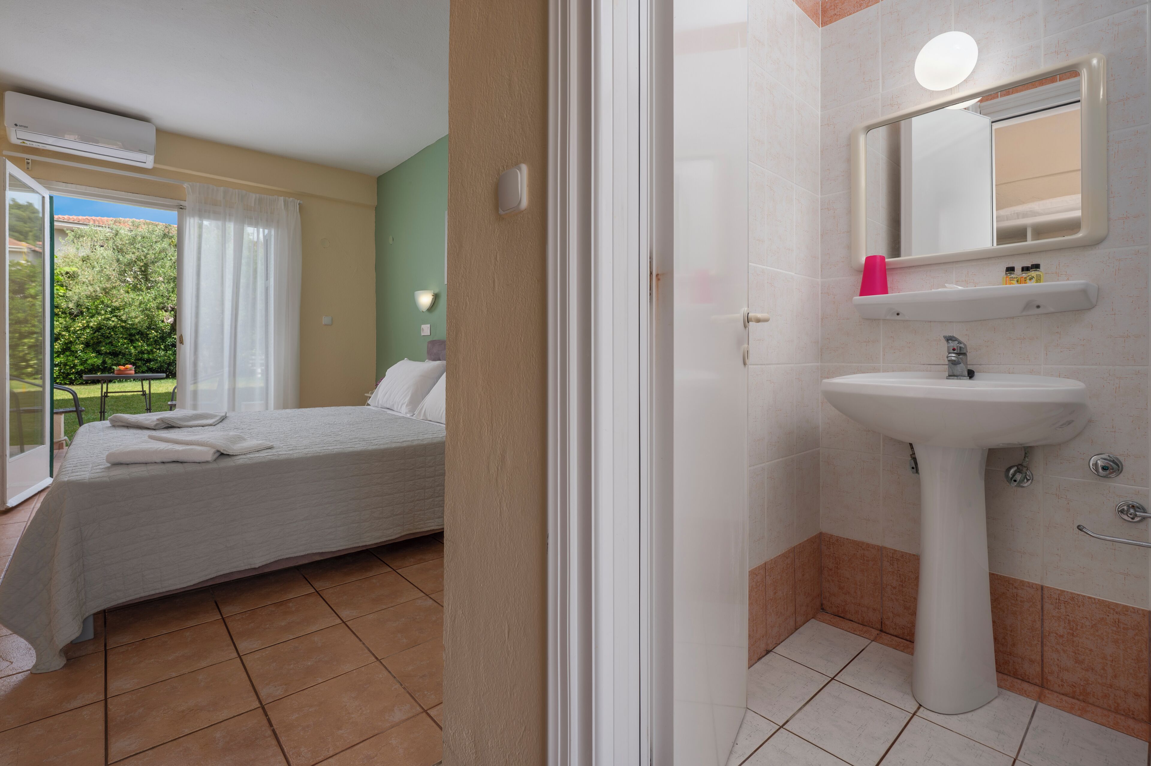 Une chambre avec un lit, une salle de bain avec un lavabo et un miroir, et une vue sur la verdure à l’extérieur.