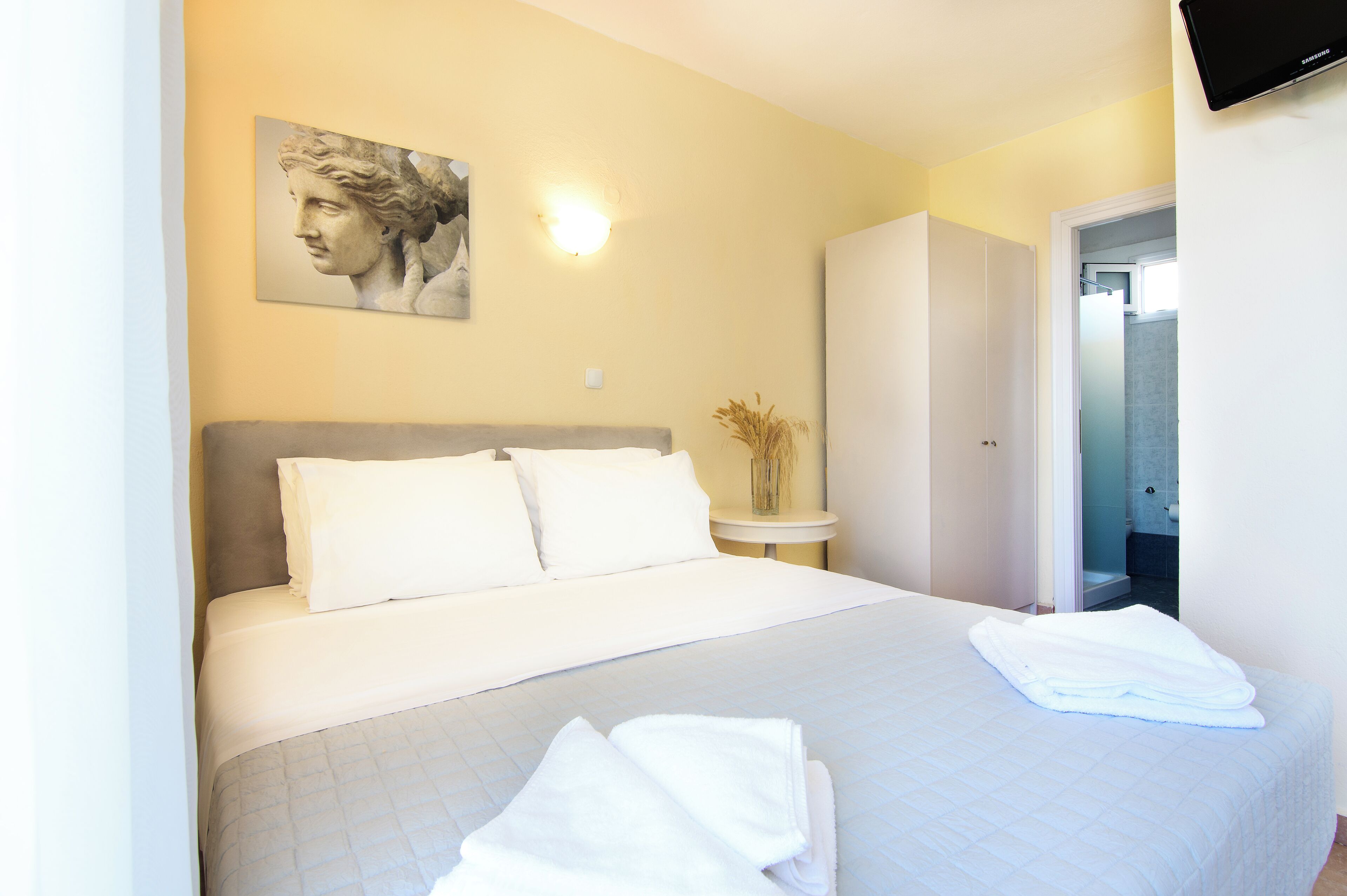 A neatly made bed with white linens, a bedside table with a vase of dried plants, and a wall-mounted light fixture.