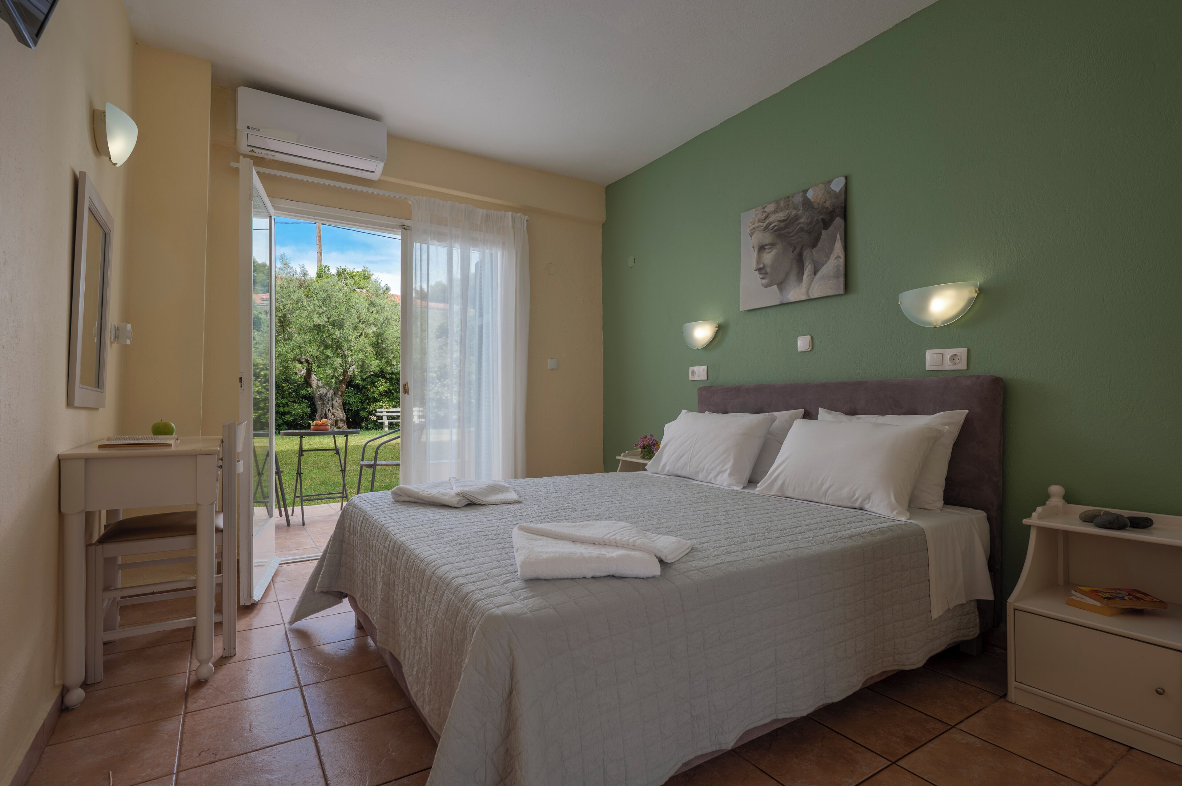 A bedroom with a bed, a nightstand, a small table, and a chair. There is a window with curtains, a painting on the wall, and an air conditioning unit.