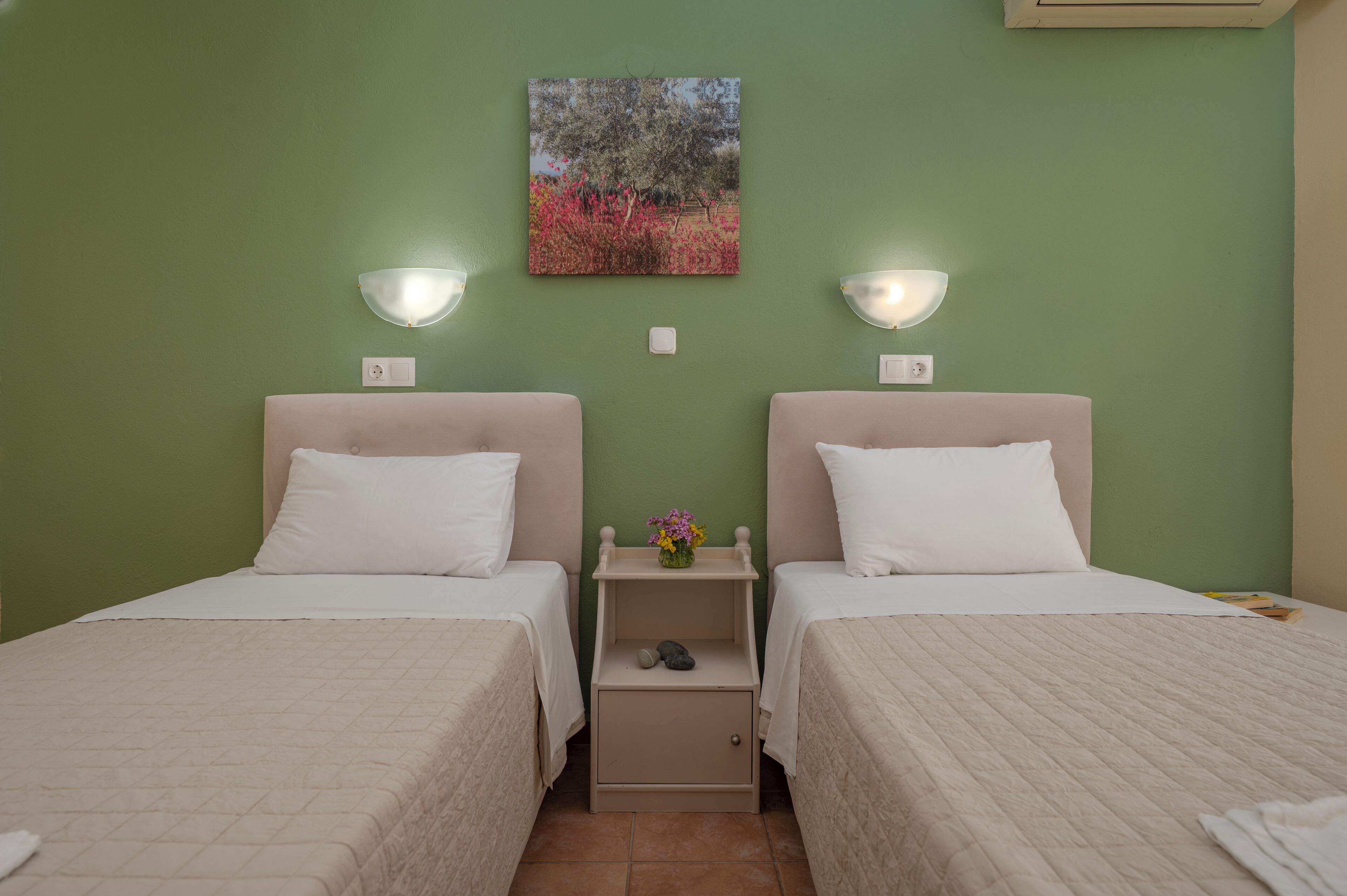 Una habitación con dos camas, cada una con un almohadón blanco y un cabecero color beige. Hay una mesita de noche con un florero y un par de zapatos. Las paredes están pintadas de verde, y hay un cuadro sobre las camas.