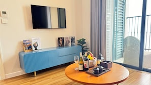 A modern living room with a round wooden coffee table, a blue sofa, a flat-screen TV mounted on the wall, and a large window with blinds.