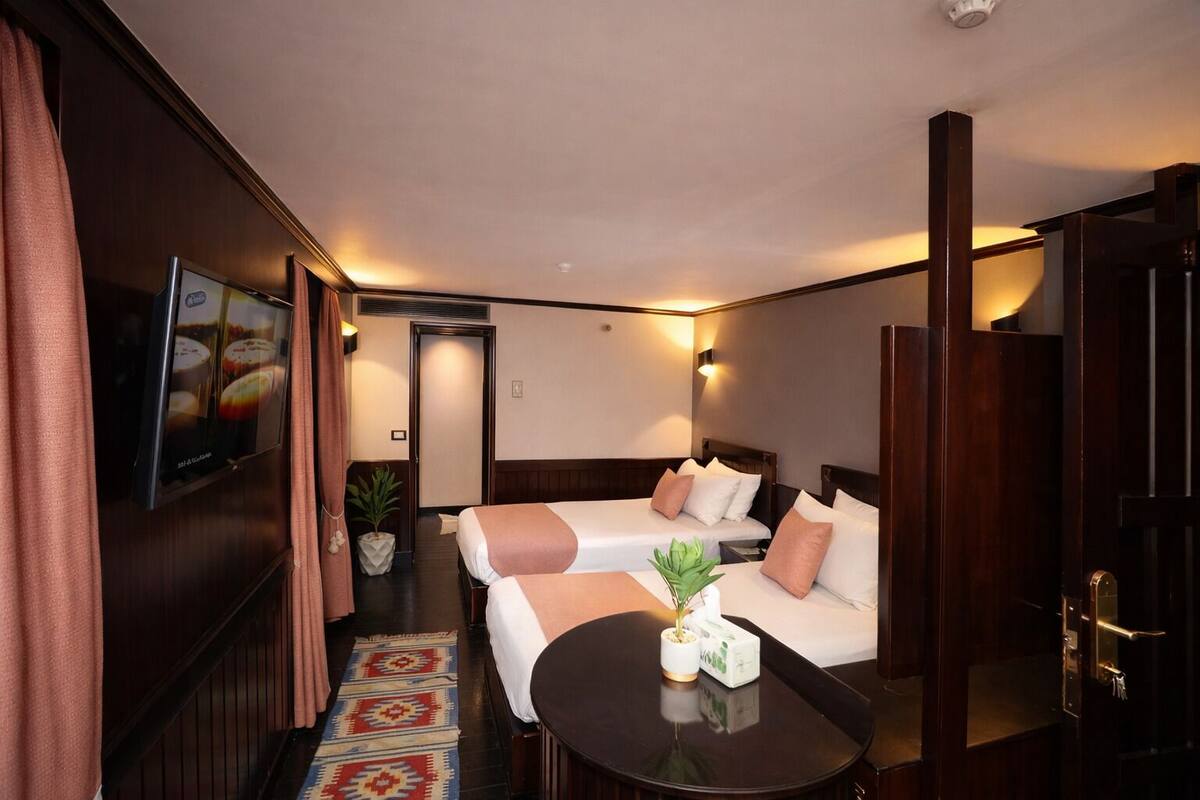 A hotel room with two beds, a small table, a potted plant, and a wall-mounted TV.