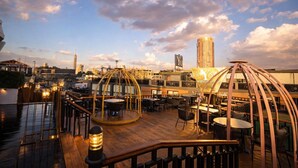 An outdoor rooftop bar with wooden flooring, round tables, and chairs. There are two large, circular, metal-framed structures providing shade. A tall building with a sign 'YU' is visible in the background. - YOU Cairo Nile View Hotel (Cairo)
