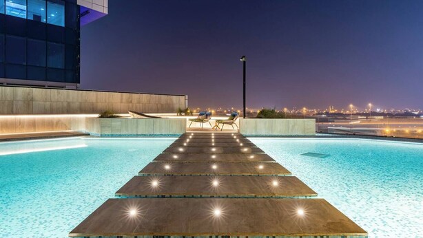 A nighttime swimming pool with a wooden deck, two lounge chairs, and a cityscape view in the background. - Bespoke Residences - Urban Oasis  (Dubai)