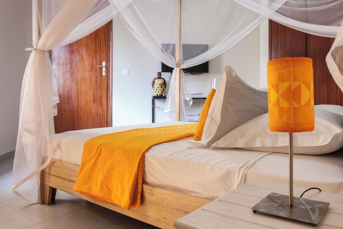 A bedroom with a wooden bed, white bedding, an orange throw blanket, a bedside table with a lamp, and a mosquito net over the bed.
