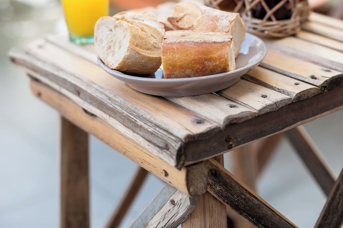 A rustic wooden table with a plate of bread and a glass of orange juice, accompanied by wicker baskets with jars, set on an outdoor patio.