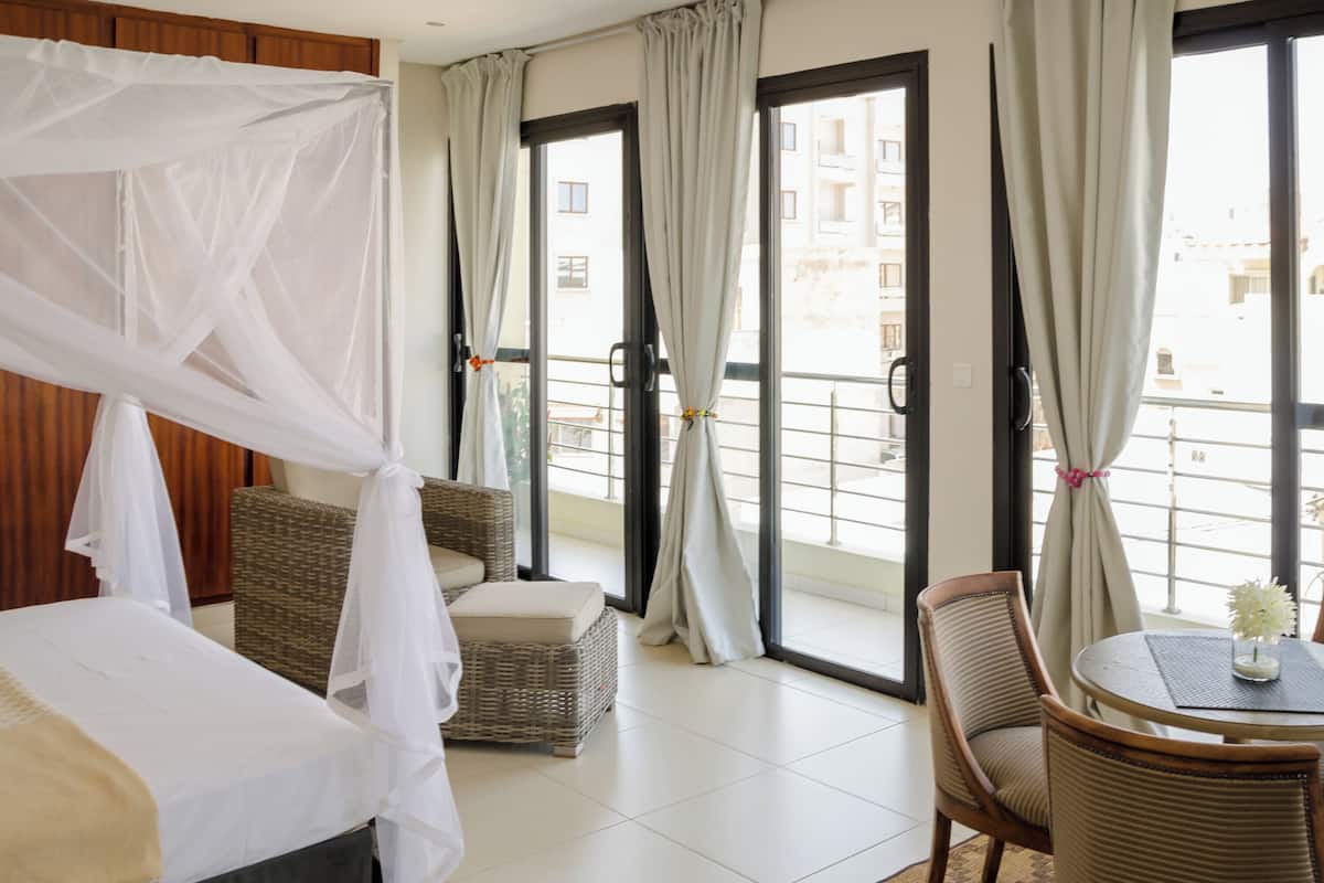 A hotel room with a bed, a wicker chair, a small table with a vase, and a balcony with curtains.