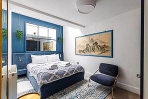 A bedroom with a blue accent wall, a bed with a patterned bedspread, a chair, a large painting, and a hanging plant.