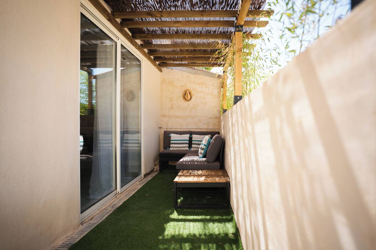 A small, enclosed patio area with a sofa and a coffee table, a pergola overhead, and a glass door leading to a bathroom.