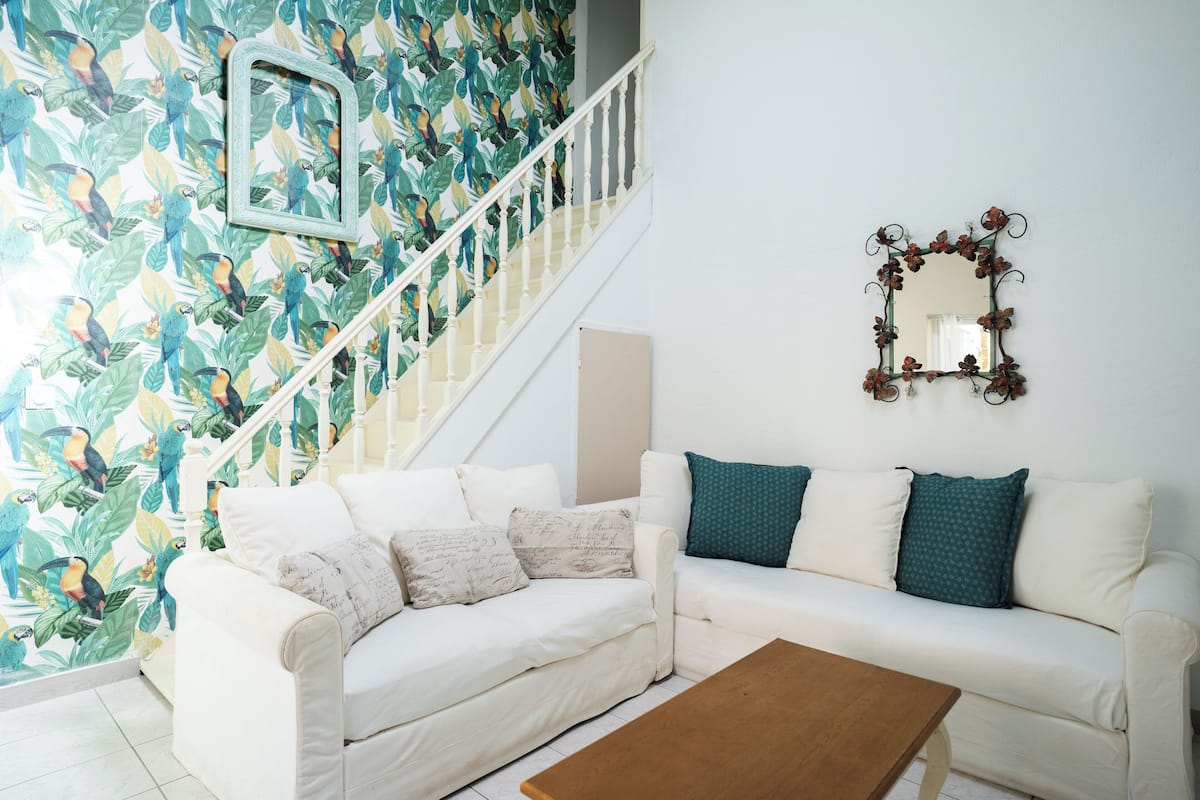 A living room with a white sofa, a wooden coffee table, a staircase with a white railing, and a wall with a mirror and a floral wallpaper.