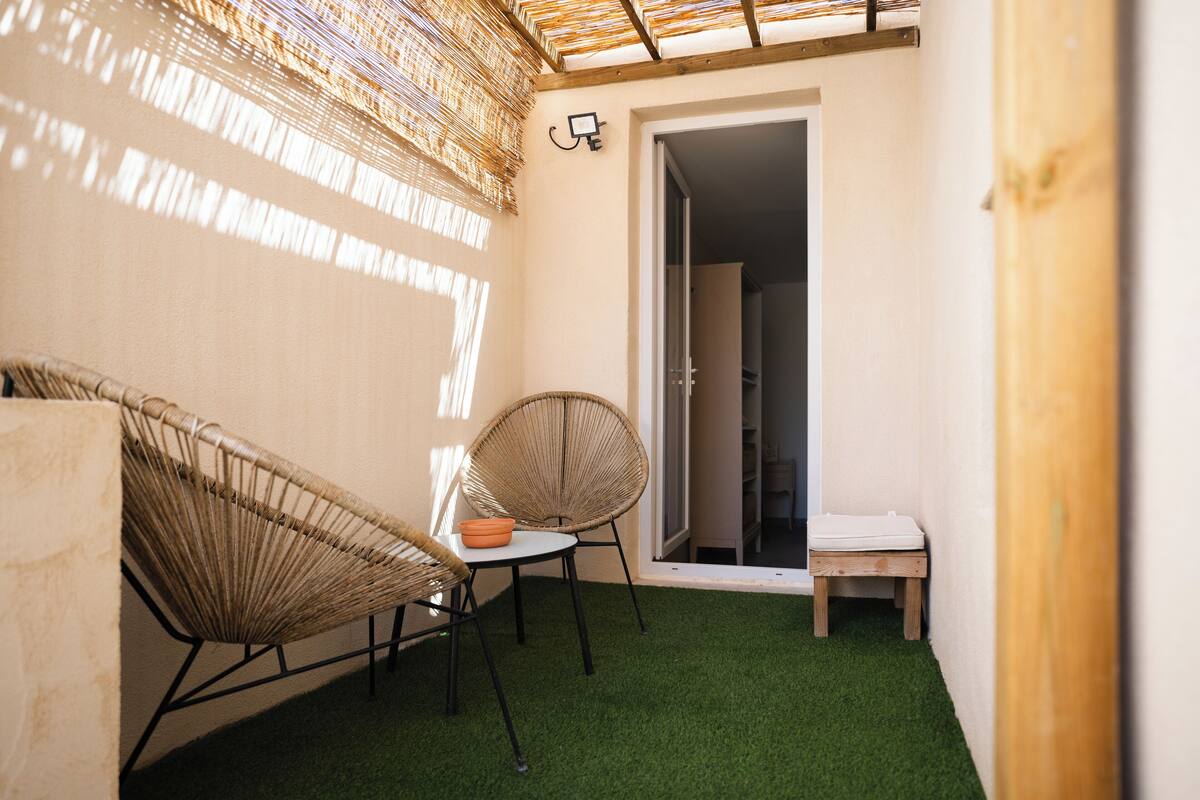 A small, enclosed outdoor space with artificial grass, wicker chairs, a small table, and a wooden bench.