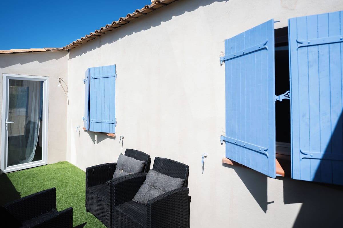 A patio with wicker furniture and blue shutters, a sliding glass door, and a white wall with a tiled roof.