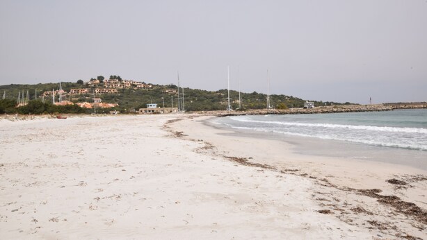 A beach with a sandy shore, a small building, and a hill with structures in the background. - Casa Ortensia (Budoni)