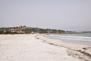 A beach with a sandy shore, a small building, and a hill with structures in the background. - Casa Ortensia (Budoni)