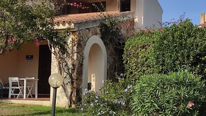 A two-story building with a stone archway entrance, a balcony with red tiles, and a garden with various plants and flowers. - Casa Ortensia (Budoni)