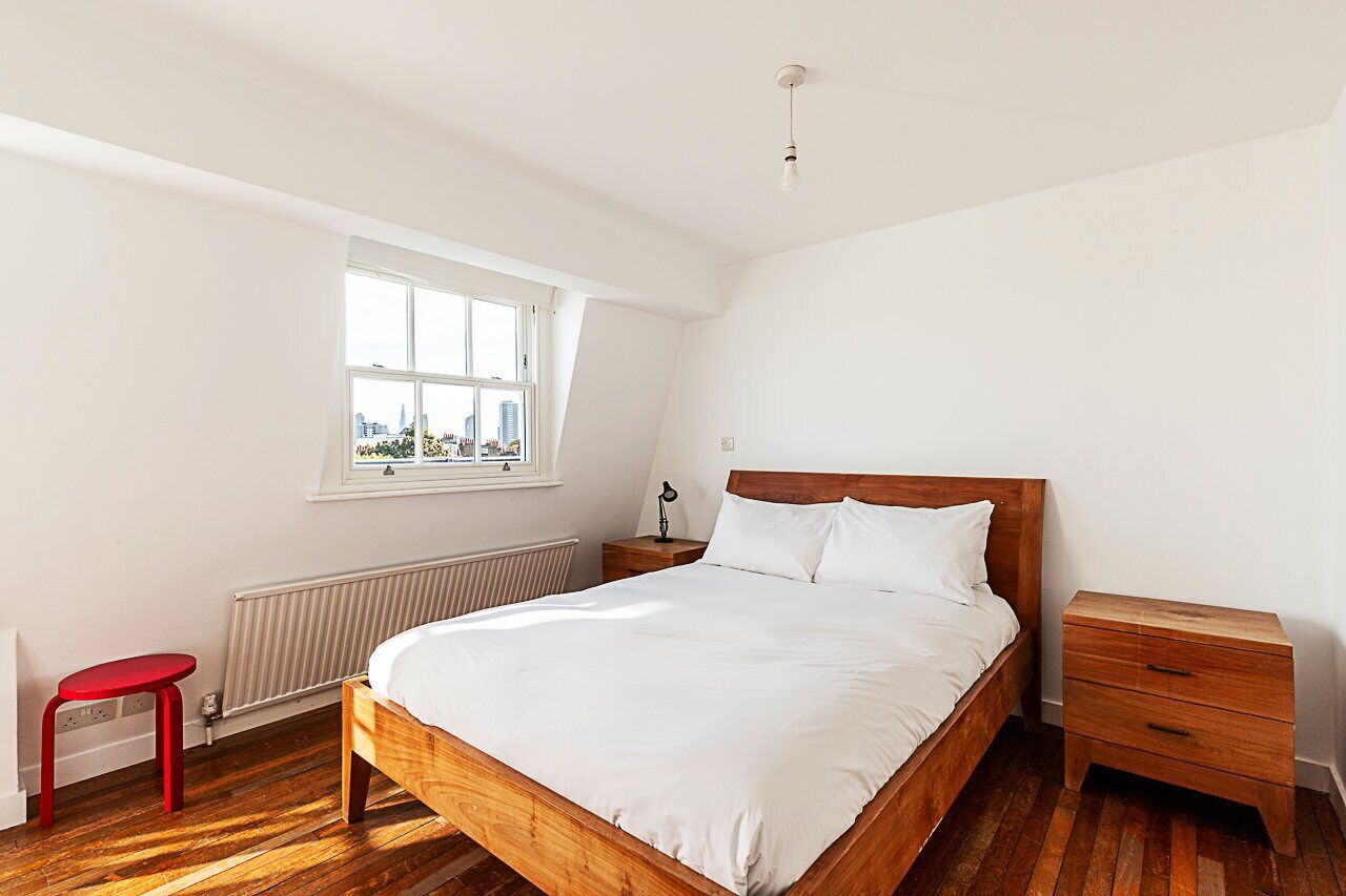Ein Schlafzimmer mit einem hölzernen Bett, einem roten Hocker, einem Nachttisch und einem Fenster mit Blick auf Gebäude.