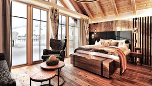 A modern bedroom with a large bed, a chair, a small table, and a view of a snowy landscape through large windows. - Hotel Salnerhof Ischgl (Ischgl)