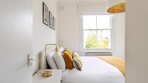 Una camera da letto con un letto, un comodino, una lampada, una finestra e un quadro appeso al muro.
